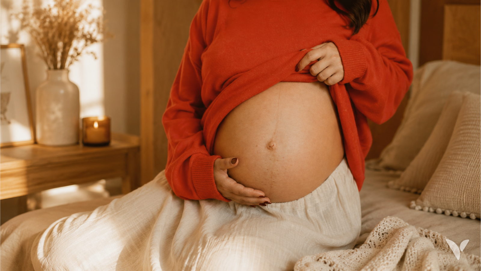 Femme enceinte assise sur un lit, une main posée sur son ventre, dans une chambre à la lumière chaude