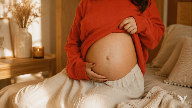 Femme enceinte assise sur un lit, une main posée sur son ventre, dans une chambre à la lumière chaude